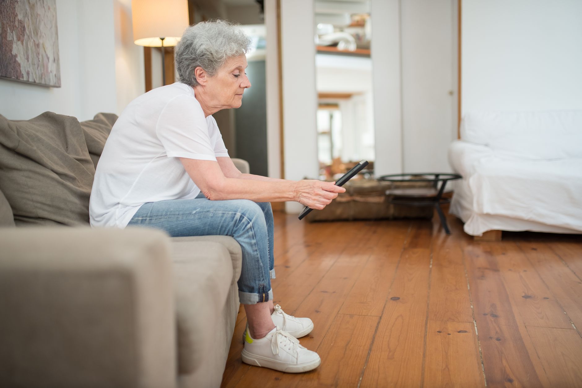 Woman in a white t-shirt and jeans sitting on a sofa and holding a tablet in a bright wooden-floored living room