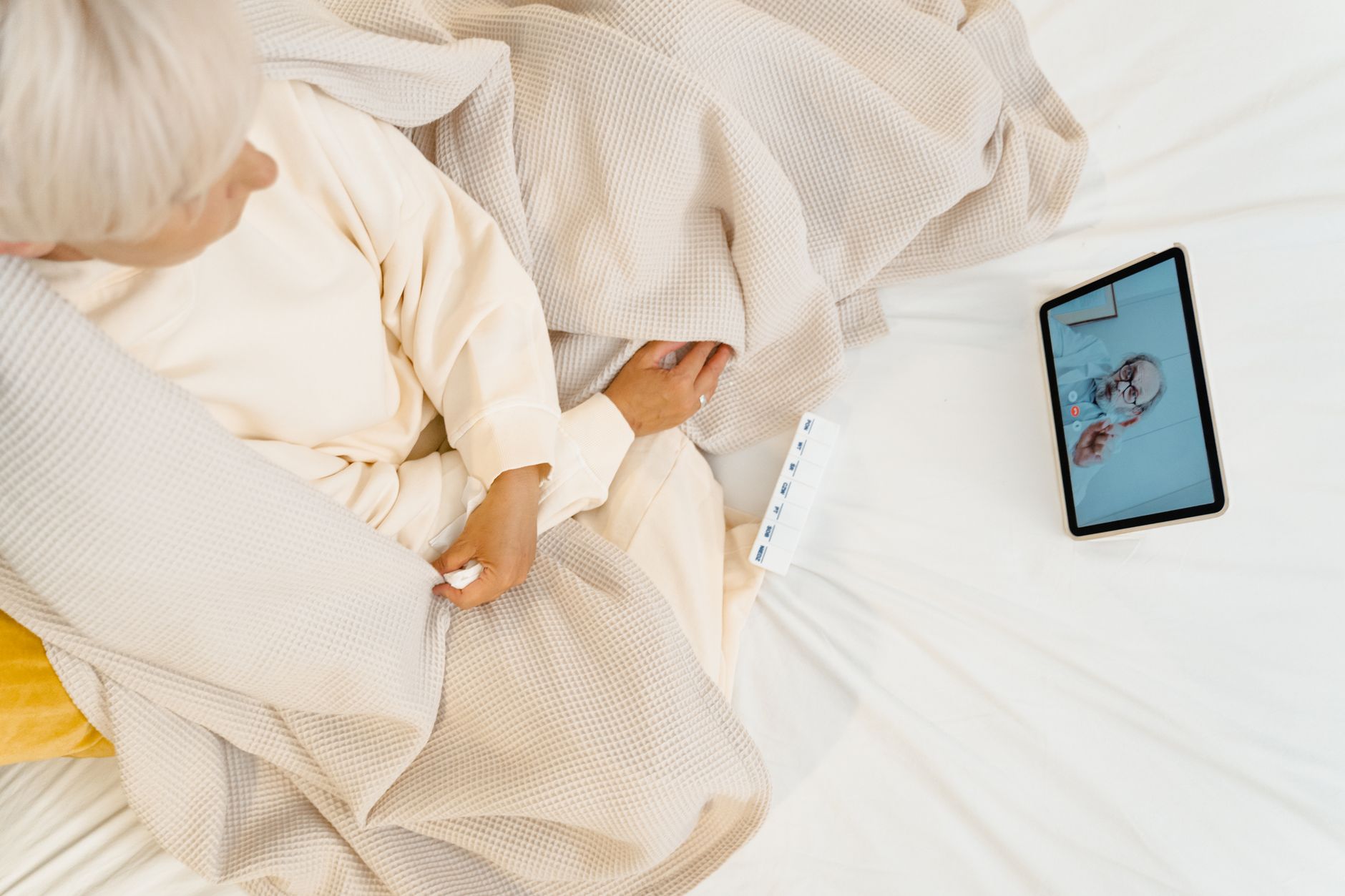 Overhead view of a person sitting on a bed with a cream blanket beside a tablet showing a video call