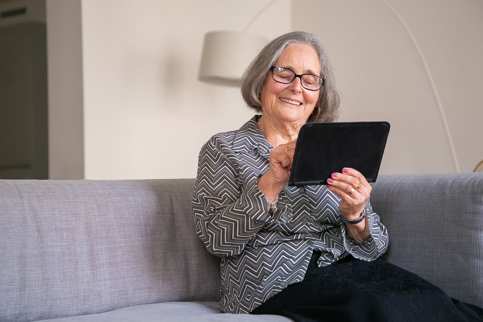 Smiling woman with glasses sitting on a grey sofa, tapping a tablet in a bright living room